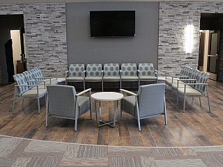Waiting area with grey chairs and chairs with a grey circular pattern. Grey brick walls and wall mounted tv behind.